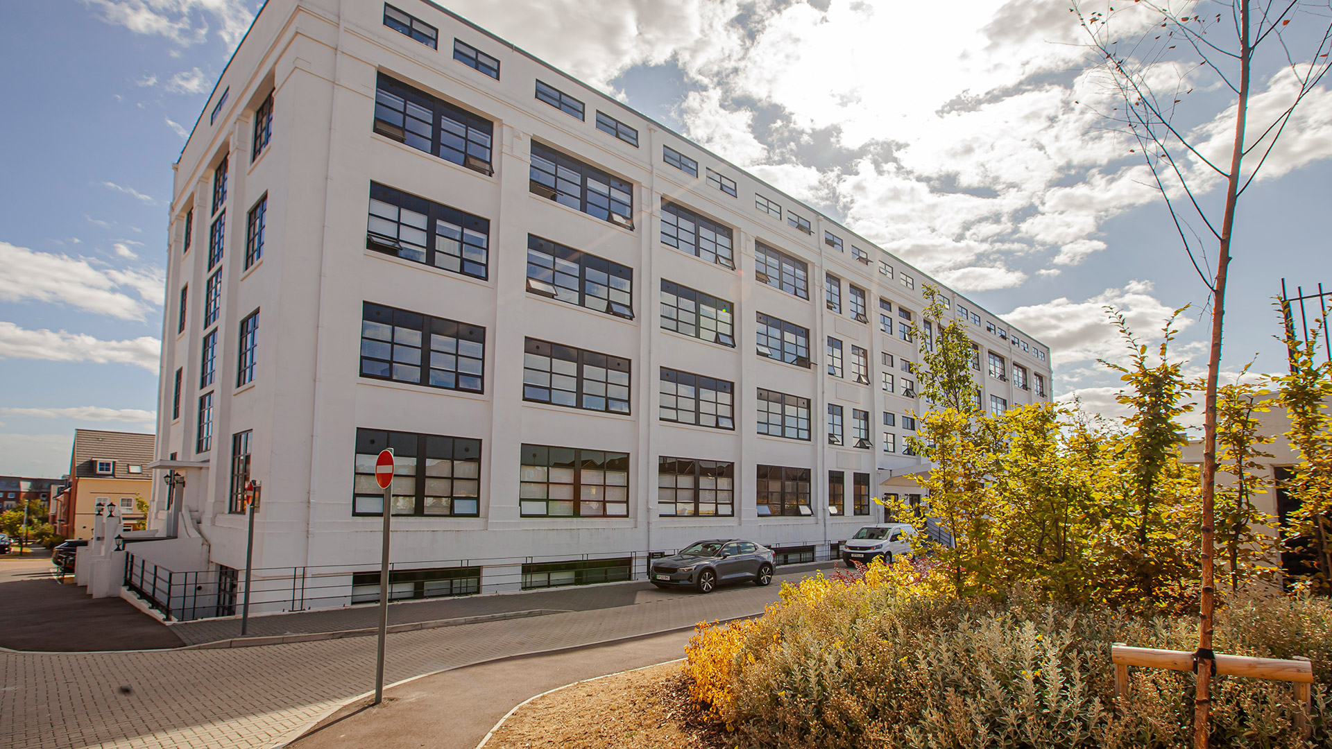 The White Building along its length: a five-storey white rendered 1930s art deco block with long horizontal bands of black Crittall-style windows and a stepped parapet, young trees and golden autumn planting in front.