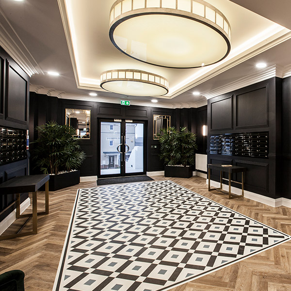 The art deco entrance lobby at The White Building: black panelled walls lined with brass post boxes, a herringbone timber floor, a geometric floor inlay, dark green velvet armchairs and potted palms.